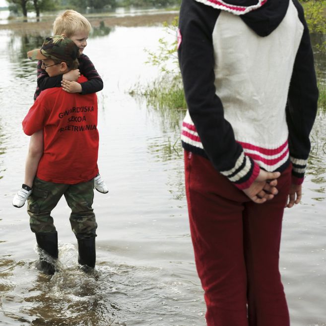 Ein Junge trägt ein kleineres Kind durch flaches Hochwasser, während eine Person im Vordergrund zuschaut.