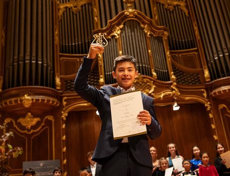 Ein junger Pianist hält stolz eine Urkunde und eine Trophäe in einem Konzertsaal mit einer großen Orgel im Hintergrund.