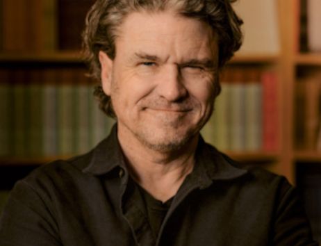 Ein Mann mit grauen Haaren und Bart lächelt vor einem Bücherregal.