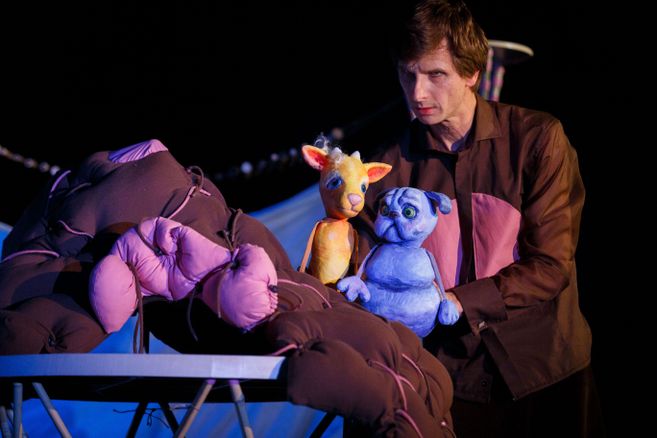 Ein Puppenspieler hält zwei bunte Puppen, eine gelbe und eine blaue, vor einem dunklen Hintergrund.