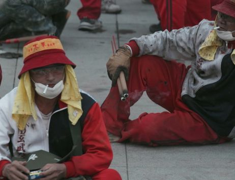Sie sehen ein Filmstill aus Yalda Afsahs Arbeit HEAT: Zwei Männer sitzen auf einem Steinboden. Sie tragen rot-weiße Kleidung, Hüte mit taiwanischem Schriftzug, Schutzbrillen und Mundschutz. Beide rauchen, einer hält Feuerwerkskörper in der Hand.