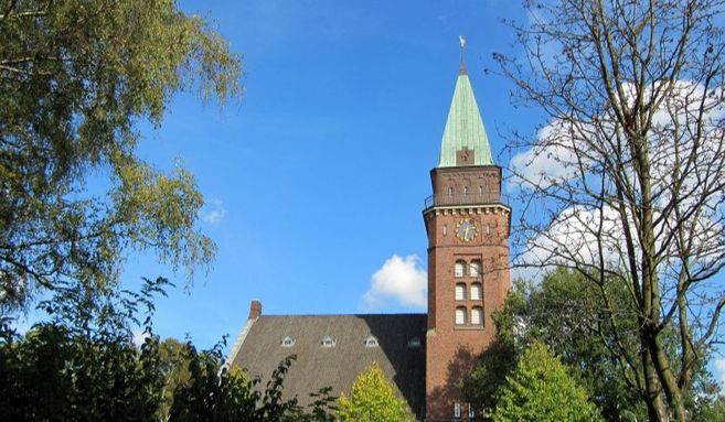 Ein Kirchturm mit grünem Dach und Uhr, umgeben von Bäumen, unter einem blauen Himmel.