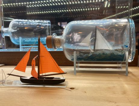 Ein Modellschiff in einer Flasche und ein kleines Modellboot mit orangefarbenen Segeln auf einem Tisch.