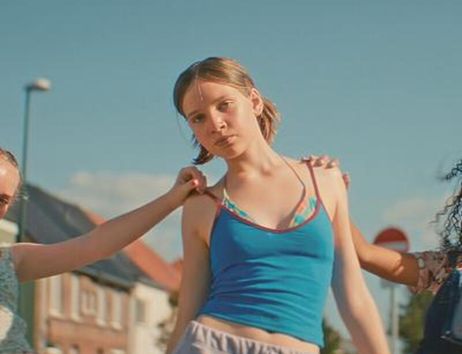 Junge Frau in blauem Top steht im Freien, während mehrere Hände sie berühren; im Hintergrund Häuser und blauer Himmel.