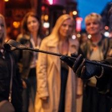 Eine Person in Lederhandschuhen hält eine Reitgerte, während vier Frauen im Hintergrund stehen.