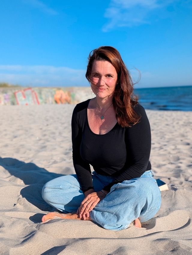 Eine Frau sitzt im Schneidersitz am Strand mit blauem Himmel im Hintergrund.