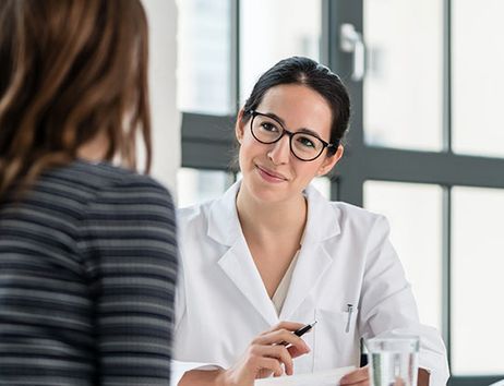 Es sind zwei Frauen zu sehen, eine sitzt vorne im Bild und ist von hinten zu sehen und eine sitzt in einem Hemd oder Arztkittel der anderen Frau gegenüber.