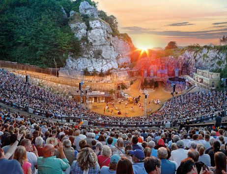 A large crowd is seated in an outdoor theater, surrounded by rocky landscapes. The sun is setting in the background, casting a warm light over the scene.