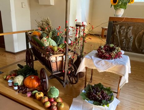 Ein Erntealtar mit einem Holzwagen voller Gemüse und Obst, umgeben von herbstlichen Dekorationen und einem Tisch mit Trauben und einem Kranz.