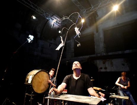 Ein Mann auf einer Bühne balanciert eine lange Stange mit Draht und Papier. Im Hintergrund sind Musiker und Instrumente zu sehen.