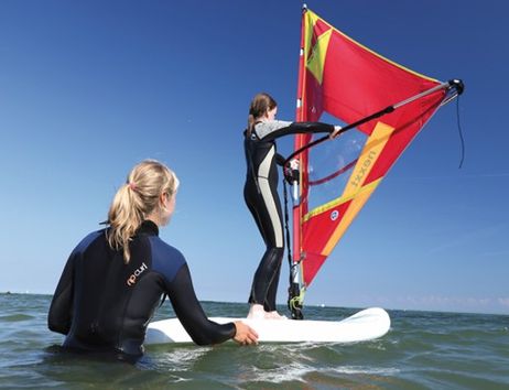 Zwei Personen beim Windsurfunterricht im Meer, eine steht auf dem Brett, die andere hält es fest.