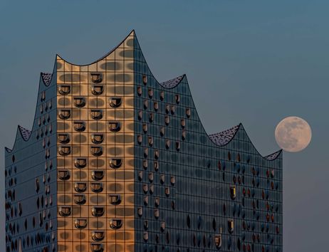 Das Foto zeigt die Elbphilharmonie in Hamburg im Abendlicht, auf deren äußerster Dachzacke ein Vollmond balanciert.