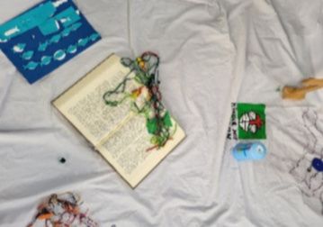Ein aufgeschlagenes Buch liegt auf einem weißen Tuch, umgeben von bunten Objekten und Zeichnungen.