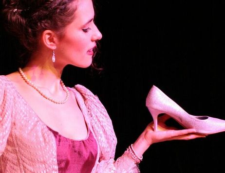 Eine junge Frau im Kostüm hält einen glitzernden rosa High Heel in der Hand, passend zum Märchen-Musical Aschenputtel.