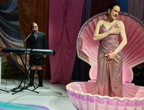 Bühnensituation: Less Brandes-Hell steht in einer rosafarbenen aufgeklappten Muschel. Sie trägt ein rosa Glitzerkleid, hält sich eine Hand vor die Brust und eine Hand vor den Schritt. Sie hat den Mund leicht geöffnet und schaut nach rechts. Im Hintergrund steht Micha Krause hinter einem Keyboard und am Mikro. Sie trägt einen schwarzen Anzug mit einem blauen Oberteil darunter sowie schwarze Stiefel. Der Hintergrund ist mit bunten Stoffen verhangen.