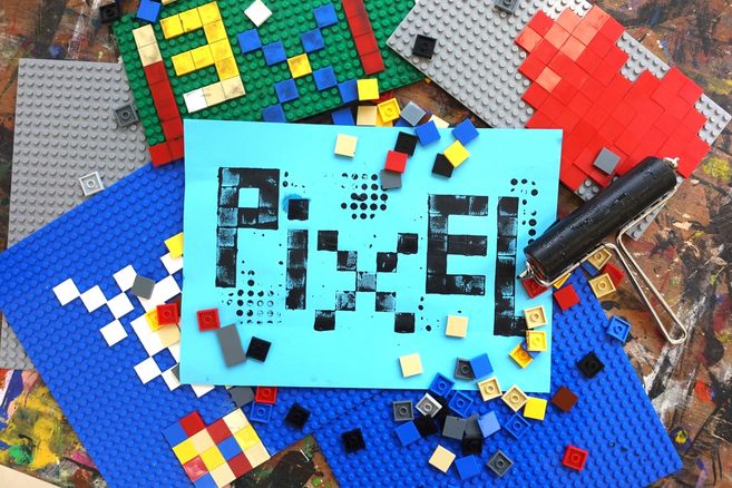 Bunte Steckbausteinplatten und lose Steine umgeben ein blaues Blatt mit dem gedruckten Wort „PIXEL“; rechts liegt eine Farbrolle.