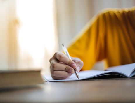 Person in gelbem Shirt schreibt mit einem Stift in ein aufgeschlagenes Notizbuch am Tisch, links liegt ein Buch im Vordergrund.