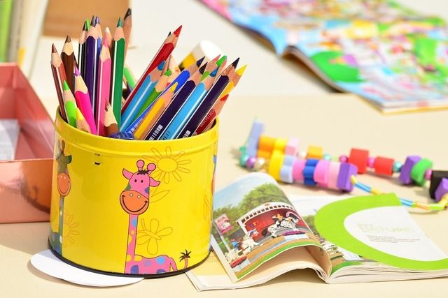 Ein gelber Behälter mit bunten Buntstiften, ein offenes Kinderbuch und bunte Perlen auf einem Tisch.